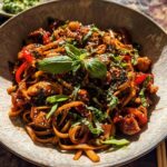 A close-up of a bowl filled with delicious Sticky Garlic Chicken Noodles, garnished with fresh herbs and sesame seeds.