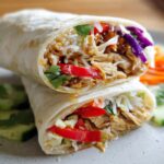 A close-up of a Teriyaki Chicken Wrap cut in half, revealing shredded chicken, rice, and colorful vegetables.