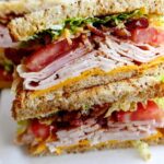 A tall, stacked Breakfast Club Sandwich with layers of turkey, bacon, tomato, lettuce, and cheese on toasted bread.