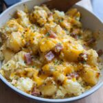 A close-up of a Cheesy Potato Egg Scramble in a pan, topped with melted cheddar cheese and herbs.