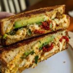 A close-up of a perfectly grilled CHICKEN AVOCADO MELT SANDWICH, revealing layers of chicken, avocado, and sun-dried tomatoes.