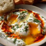 Close-up of Çılbır: Turkish eggs poached in creamy yogurt, drizzled with spiced butter and fresh herbs, served with bread.