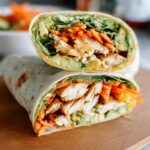 A delicious wrap filled with chicken, shredded carrots, avocado, and greens, cut in half to show the creamy spinach and eggs filling.