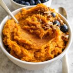 A close-up of a Delicious Sweet Potato Breakfast Bowl topped with blueberries and granola, with a spoon in the bowl.