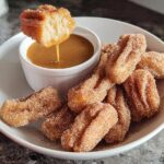 A piece of fluffy air fryer churro bite being dipped into a caramel sauce, with a plate full of churro bites.