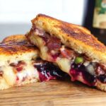 A gourmet blueberry grilled cheese sandwich, cut in half, showing melted cheese, blueberries, and bacon.