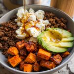 A close-up of a Ground Beef Hot Honey Bowl featuring seasoned ground beef, scrambled eggs, roasted sweet potatoes, and sliced avocado, drizzled with honey.