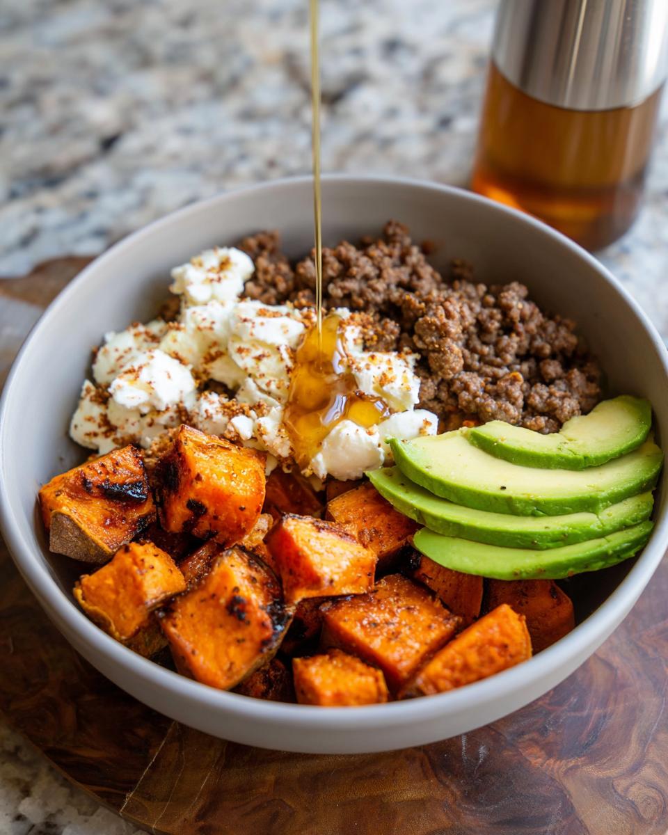 A close-up of a Ground Beef Hot Honey Bowl with roasted sweet potatoes, crumbled cheese, sliced avocado, and honey being drizzled.