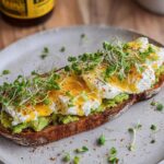 A slice of toast topped with mashed avocado, cottage cheese, poached eggs, and sprouts, a delicious high-protein breakfast.