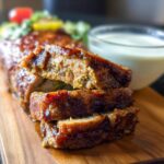 A close-up of a sliced Juicy Garlic Parmesan Chicken Meatloaf on a wooden board with a side of sauce.