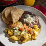 A plate of scrambled Mexican Eggs topped with tomatoes and cheese, served with refried beans and corn tortillas.
