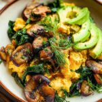 A close-up of fluffy Mushroom Spinach Scrambled Eggs topped with sliced avocado and fresh dill.