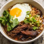 A close-up of a bowl of Slow Cooker Beef Ramen Noodles topped with a fried egg, cilantro, and green onions.