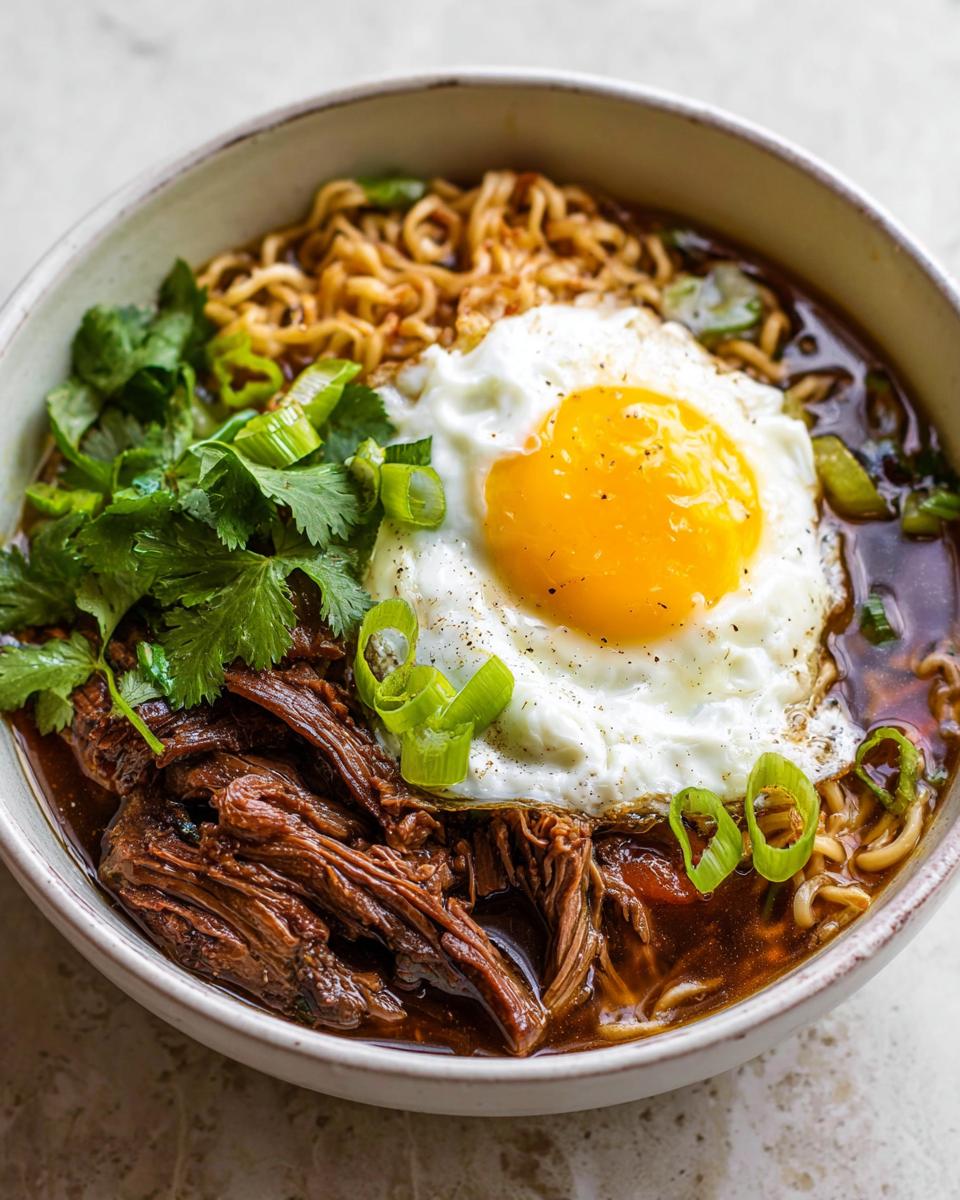 A close-up of a bowl of Slow Cooker Beef Ramen Noodles topped with a fried egg, shredded beef, cilantro, and green onions.