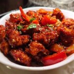 A bowl of Spicy Dragon Chicken, featuring crispy fried chicken pieces coated in a glossy, spicy sauce with red bell peppers and sesame seeds.