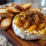 Close-up of baked brie topped with sliced peaches, toasted pecans, and herbs, served with toasted baguette slices.
