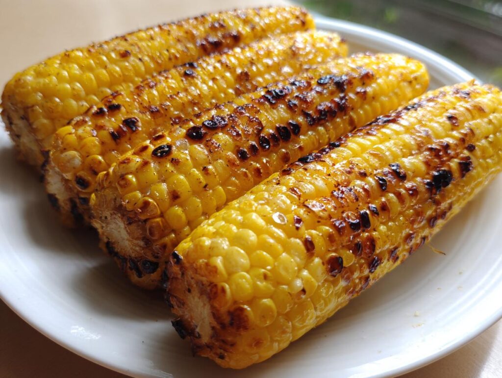 Four ears of grilled corn on the cob on a white plate, showing perfect char marks from grilling.