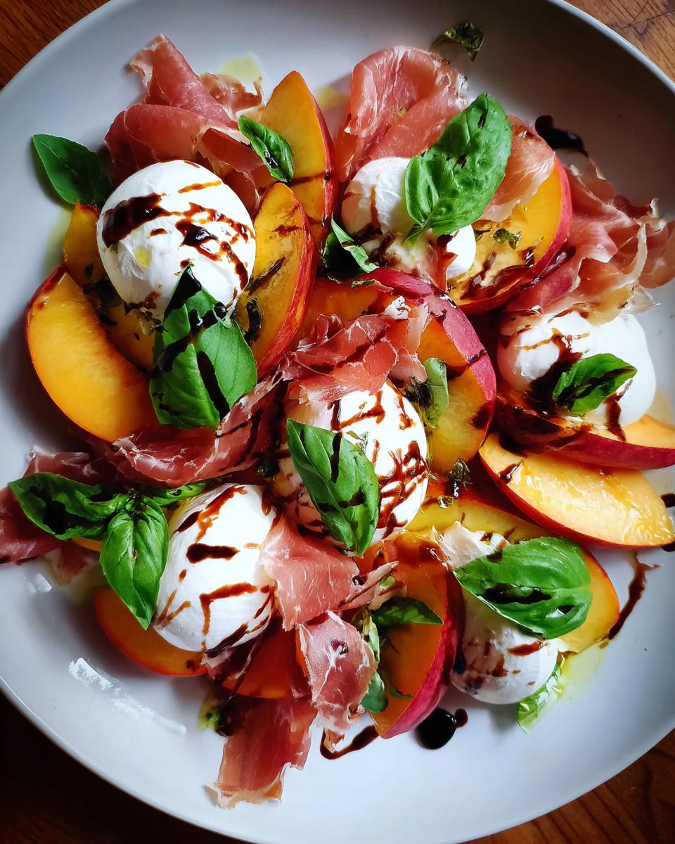 A vibrant summer salad featuring slices of fresh peaches, creamy burrata cheese, prosciutto, and basil leaves, drizzled with balsamic glaze.